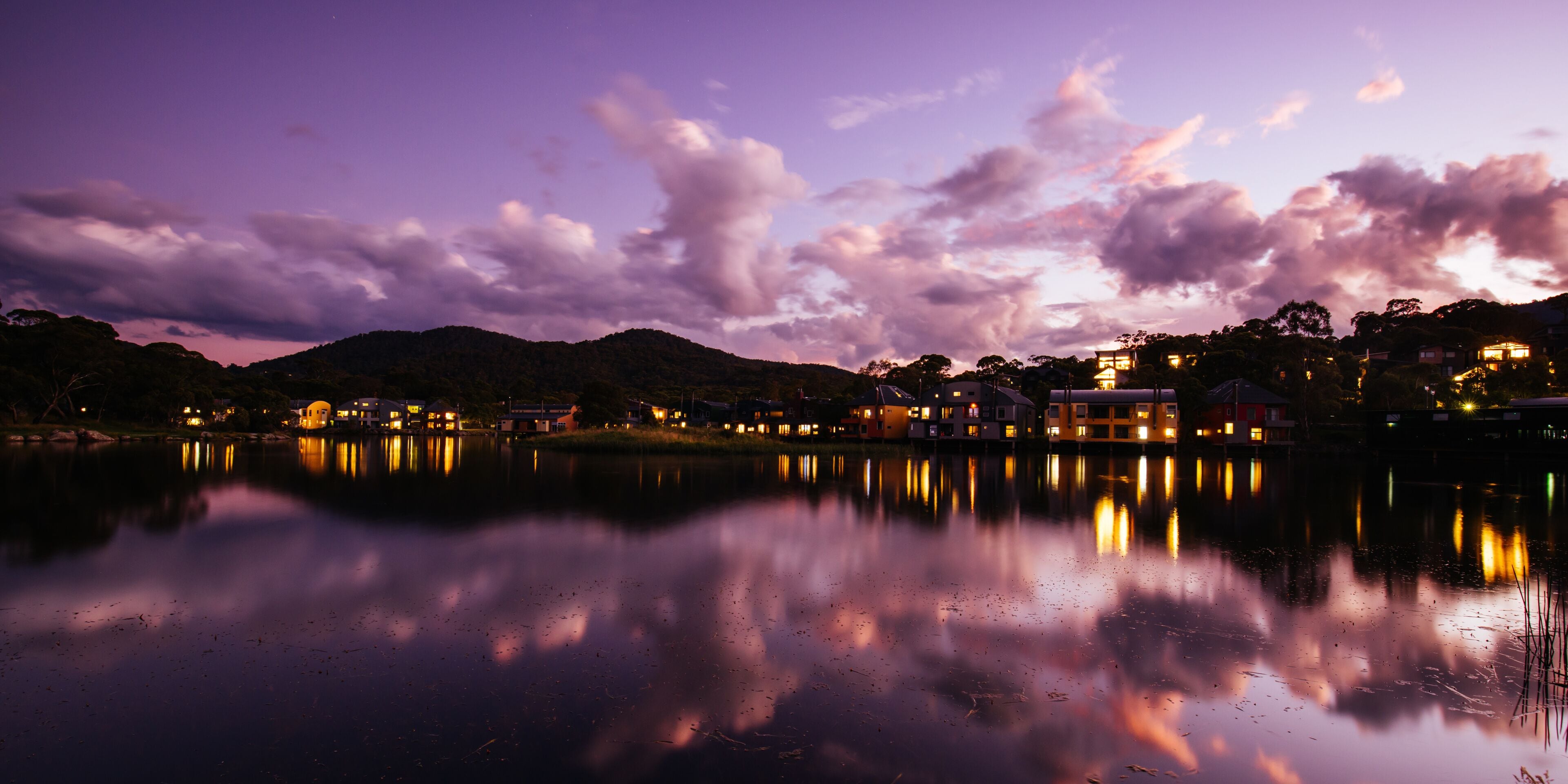 A calm lake at dusk with reflections of buildings and a mountain range in the background.