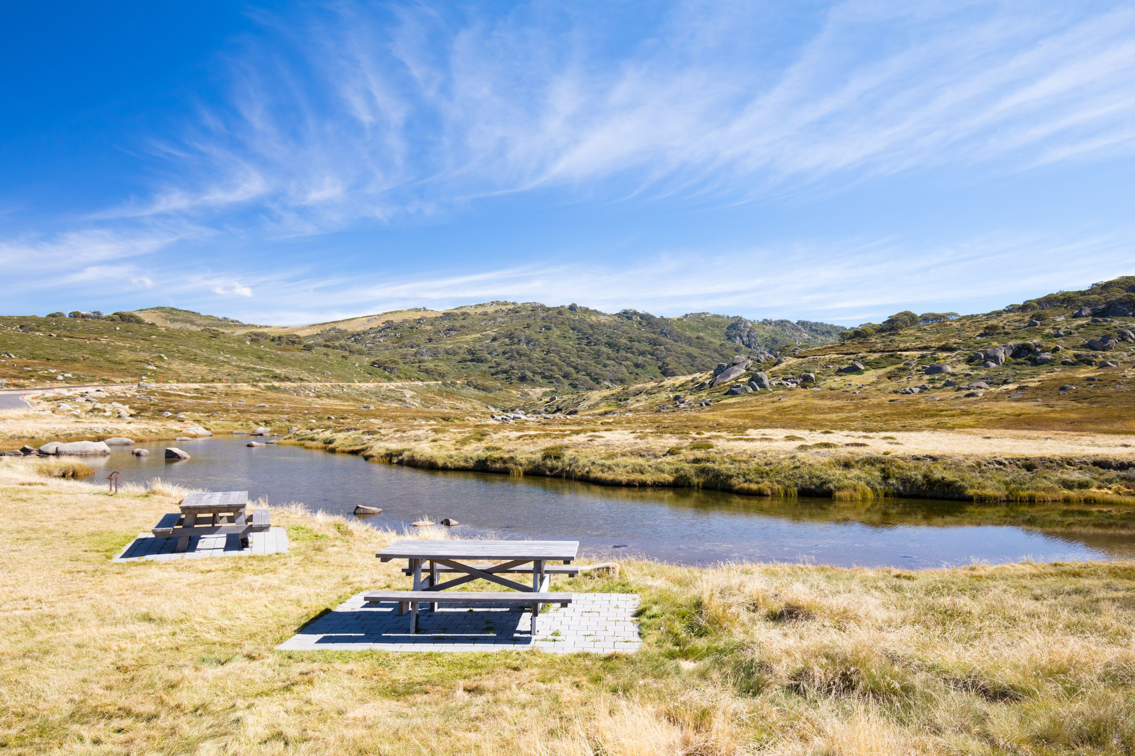 A serene lakeside picnic area with a wooden deck and benches, surrounded by rolling hills and a clear blue sky.