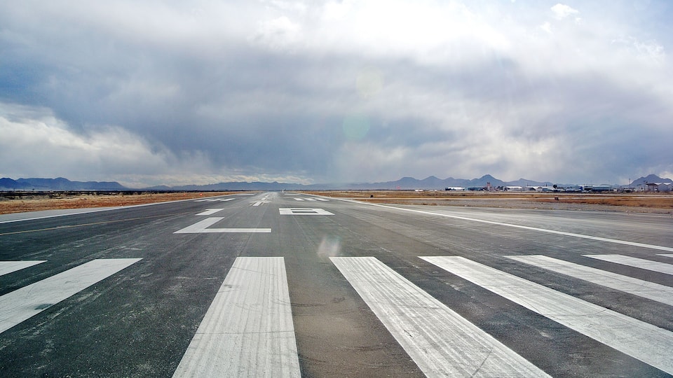 A wide runway with white stripes, surrounded by mountains and a cloudy sky.