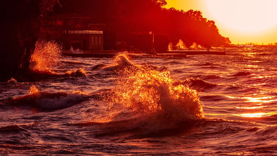 A sunset over a turbulent sea with waves crashing onto a rocky shore.