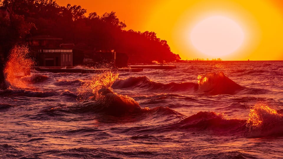 A sunset over the ocean with waves crashing onto a rocky shore.