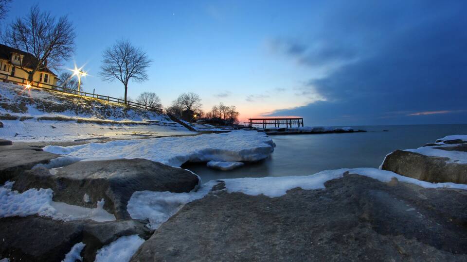 A snowy landscape with large rocks, a wooden pier, and a sunset over the water.