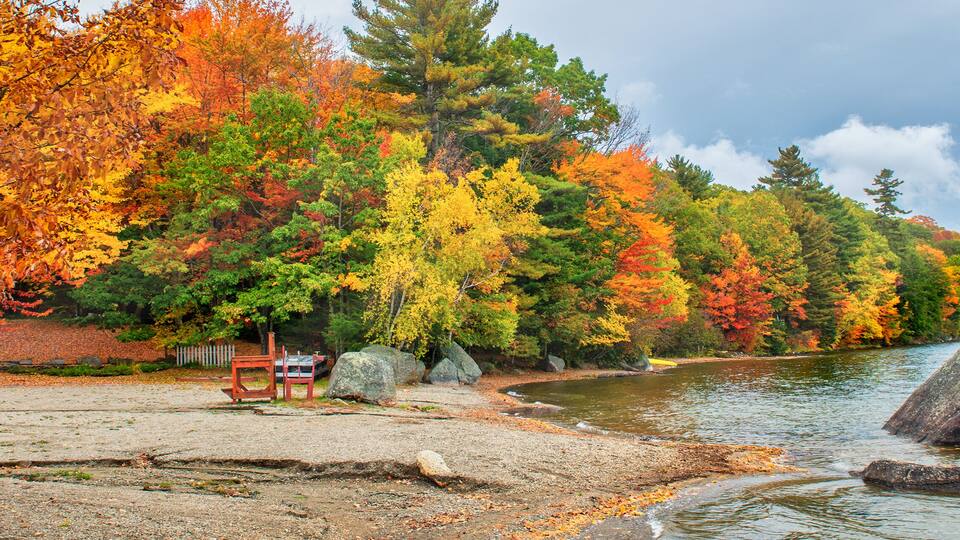 A lakeside park with autumn foliage, a wooden bench, and large rocks by the water.