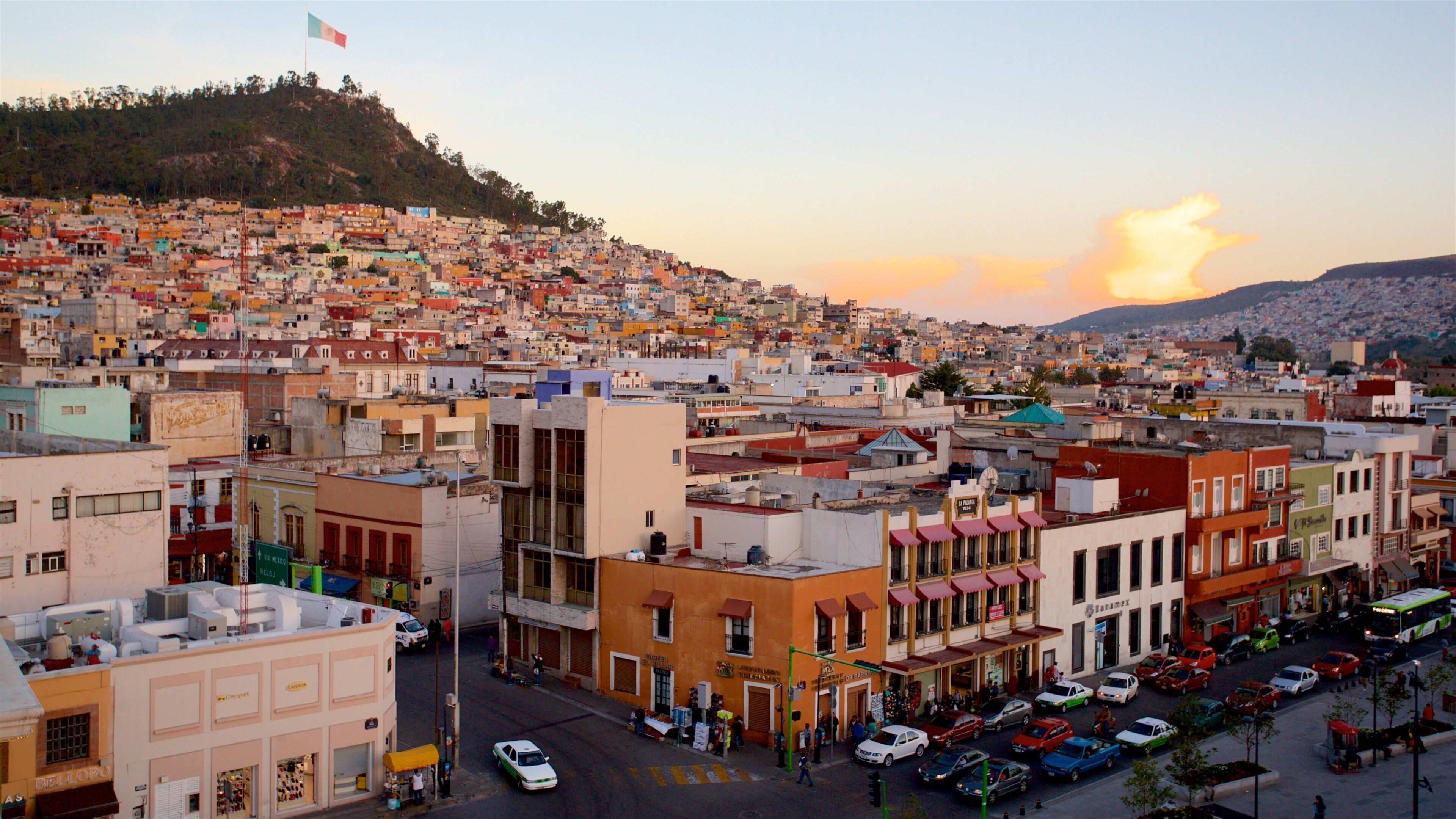 A densely packed urban area with colorful buildings, a prominent hill in the background, and a clear sky.