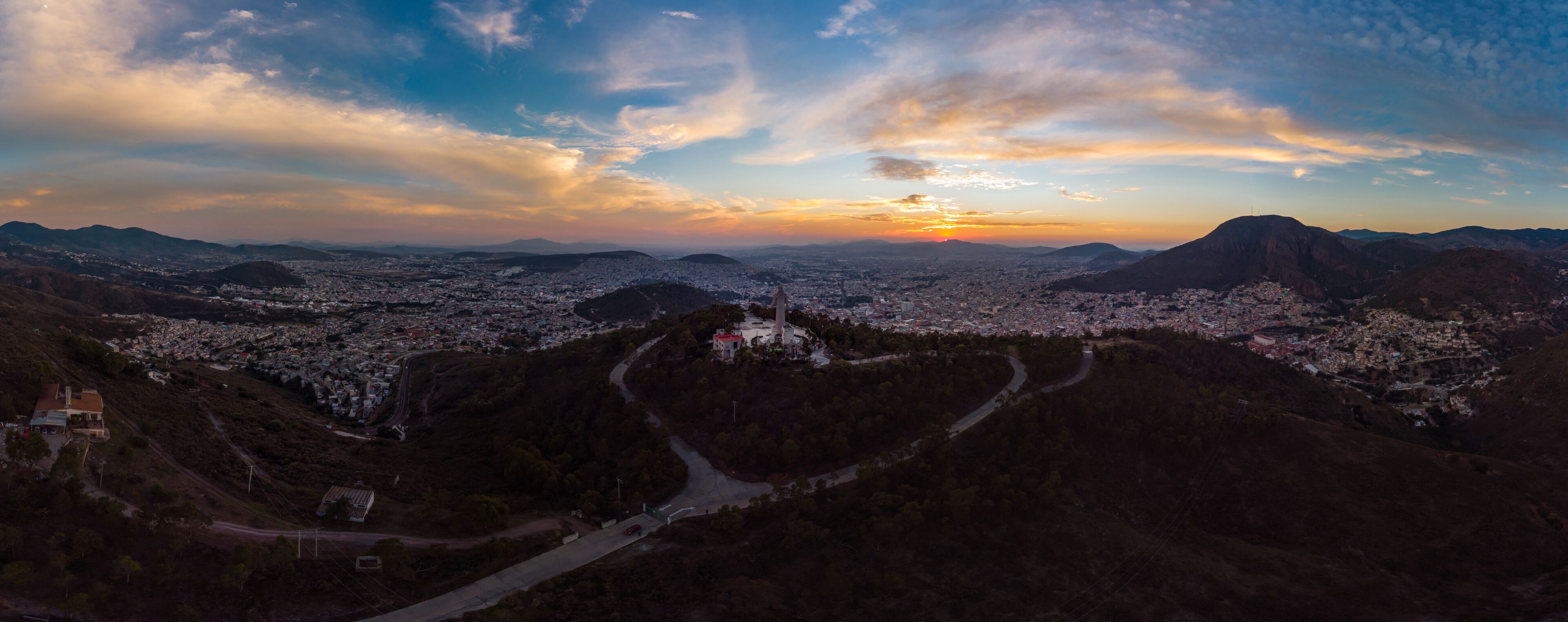 Vista panorámica de una ciudad con una colina destacada y un gran monumento en su cima, rodeada de exuberante vegetación y un camino sinuoso.
