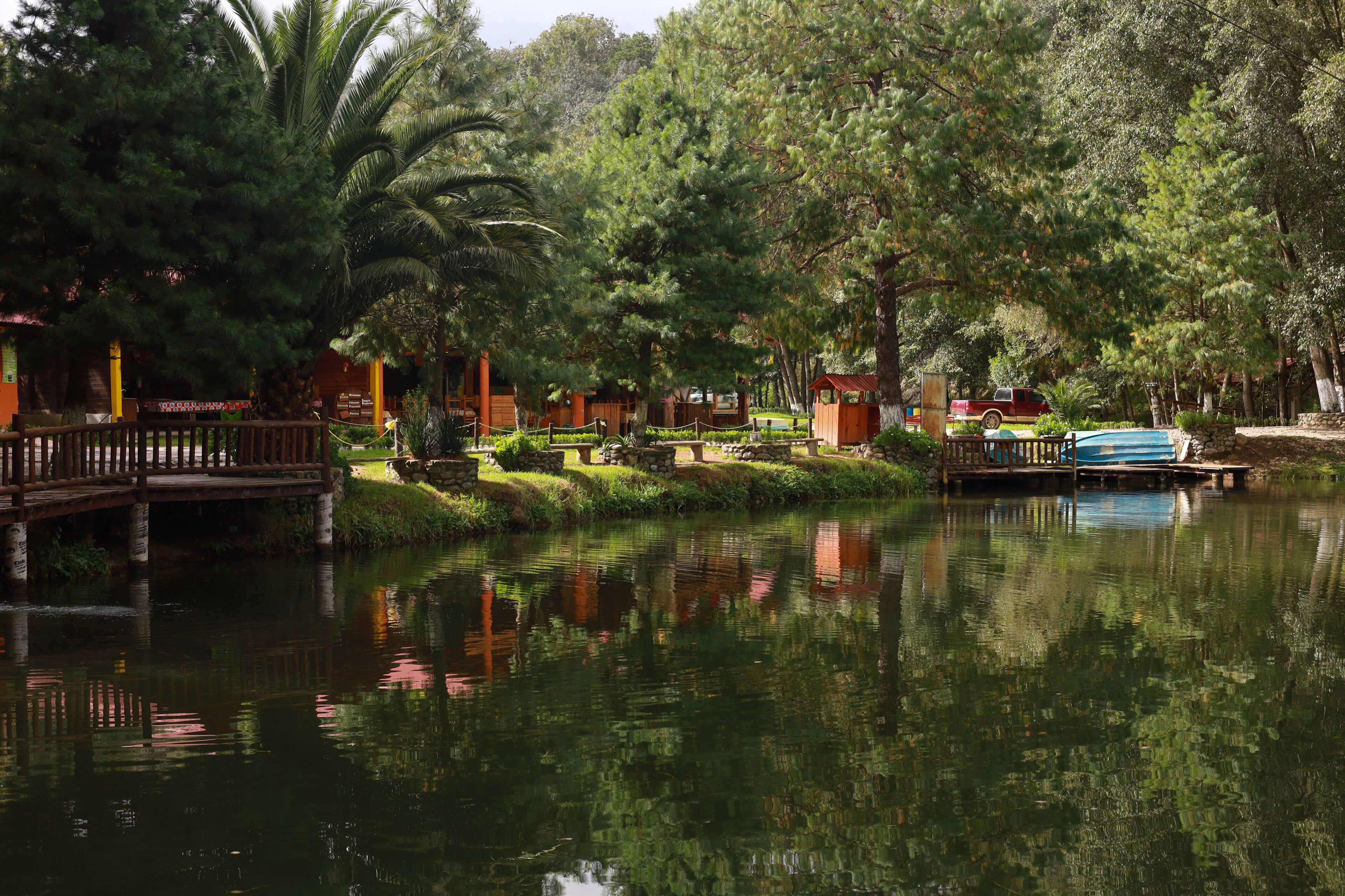 Escena tranquila junto al lago, con un puente de madera, construcciones de piedra y abundante vegetación.