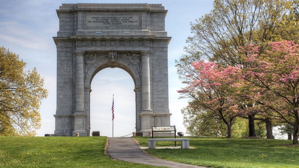A large stone archway with inscriptions, surrounded by green grass and blooming pink trees.