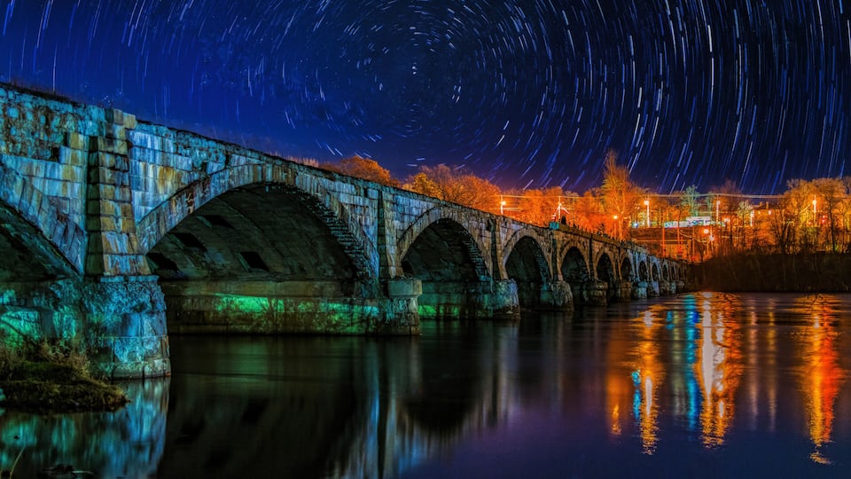 A stone bridge with star trails in the night sky above a calm river reflecting lights.