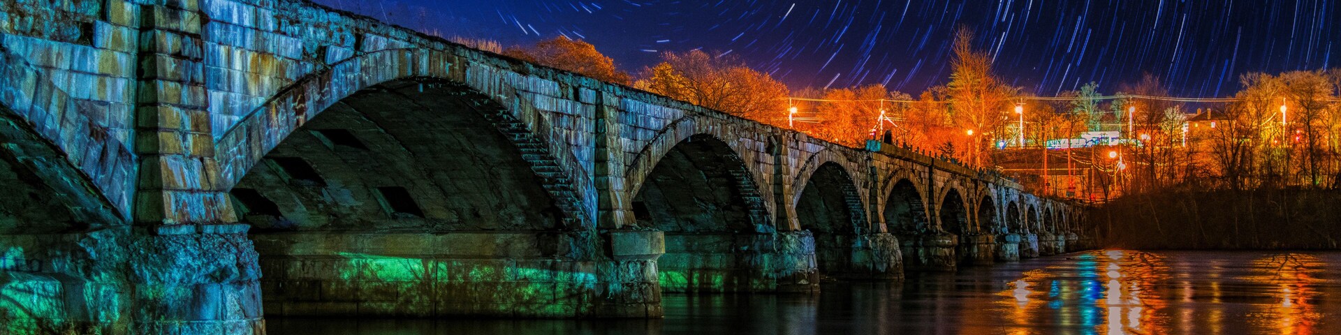 Un puente de piedra con estelas de estrellas en el cielo nocturno, sobre un río tranquilo que refleja luces.
