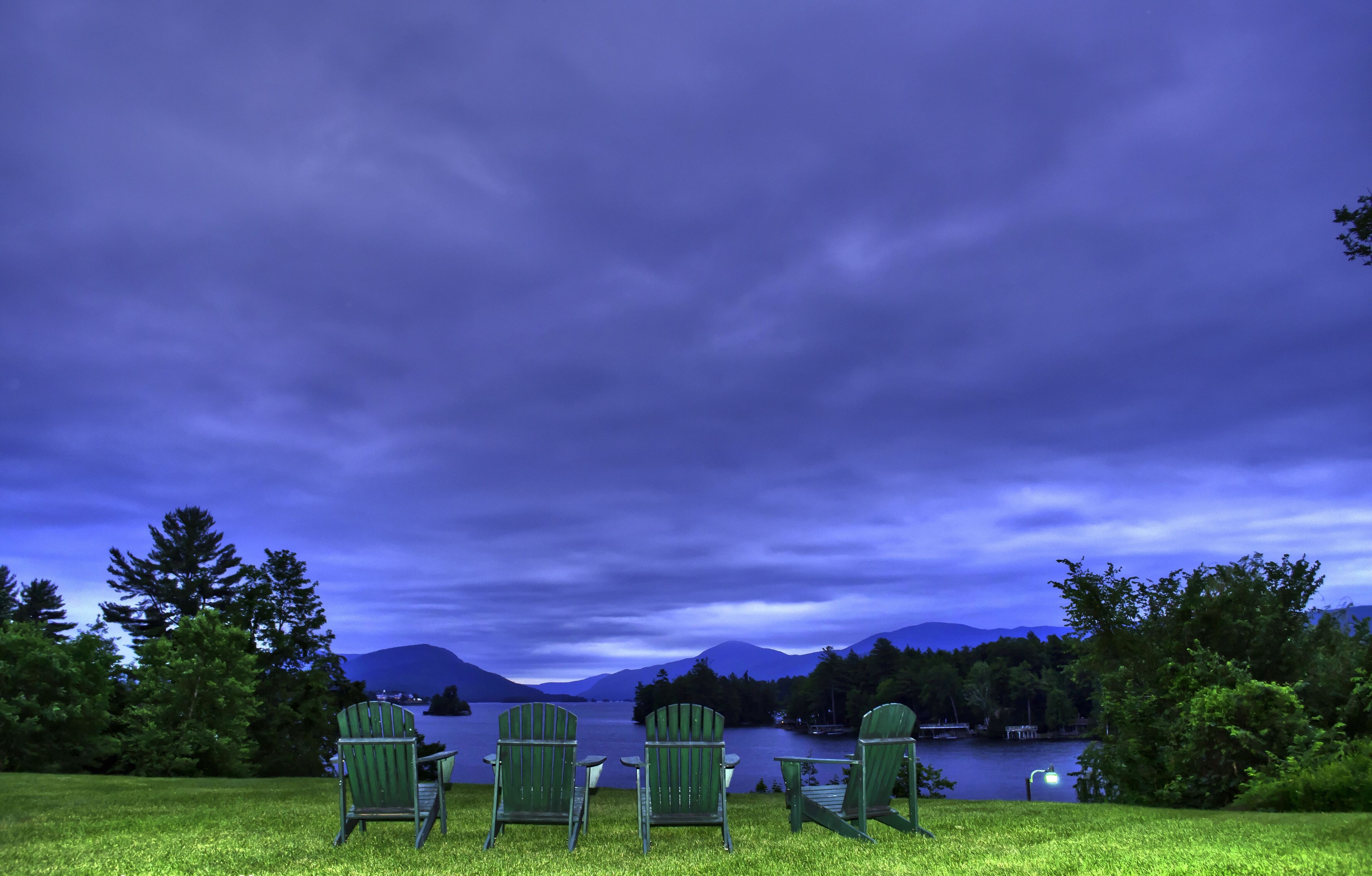 Une vue sur le lac, de nuit, avec quatre chaises Adirondack vertes disposées face à l'eau, entourées d'arbres et de montagnes, sous un ciel bleu foncé.