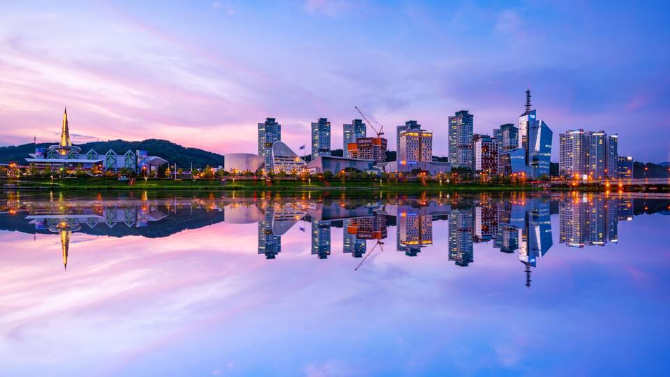 A city skyline with modern buildings and a prominent church spire reflecting on a calm body of water.