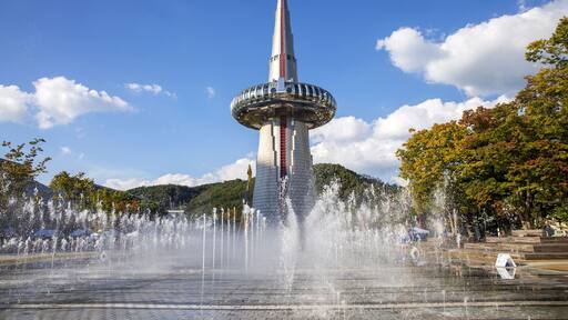 Une tour surmontée d’une flèche, entourée d’une fontaine, se dressant sur un ciel dégagé, avec des arbres en arrière-plan.