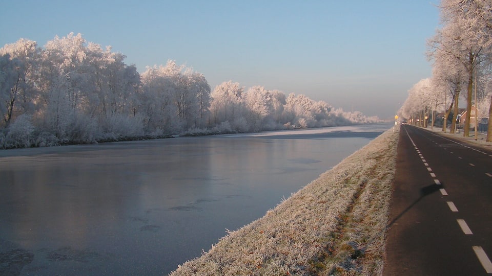 A frosty river with a road alongside, lined with trees and buildings.