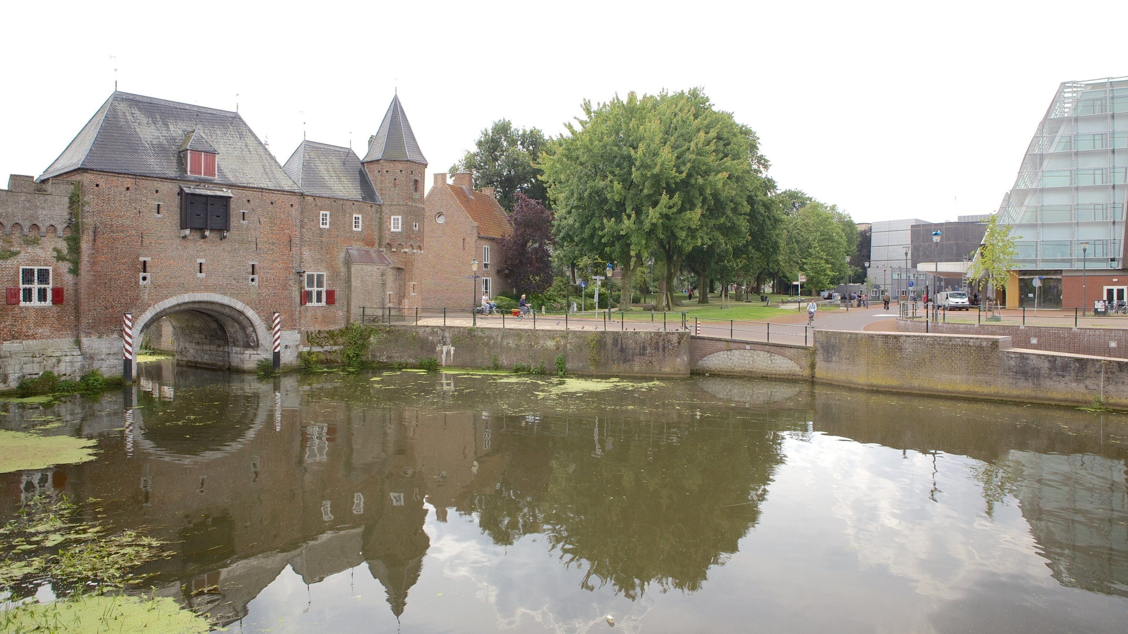 A historic brick building with a bridge over a canal, surrounded by trees and modern buildings.