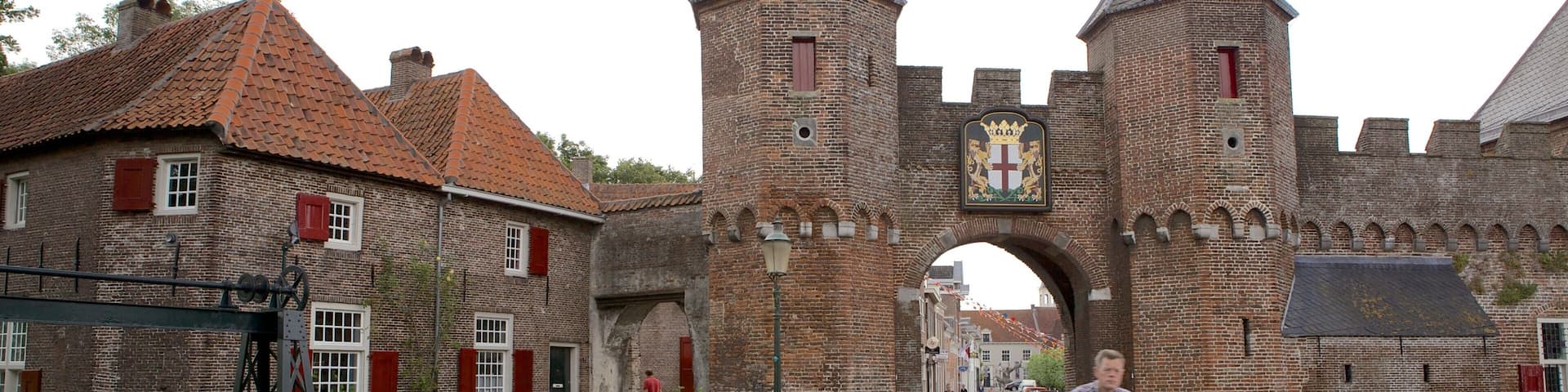 A historic brick gate with two towers, a cyclist, and traditional buildings with red shutters.