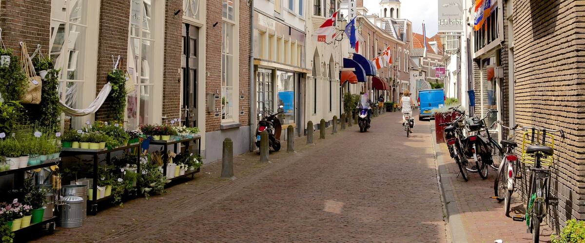 A narrow cobblestone street lined with brick buildings, a church spire in the distance, and flags hanging above.