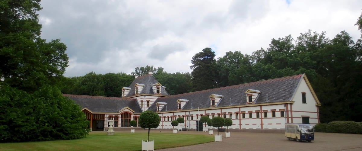 A large, historic building with a steep roof and multiple windows, surrounded by lush greenery and a well-maintained lawn.