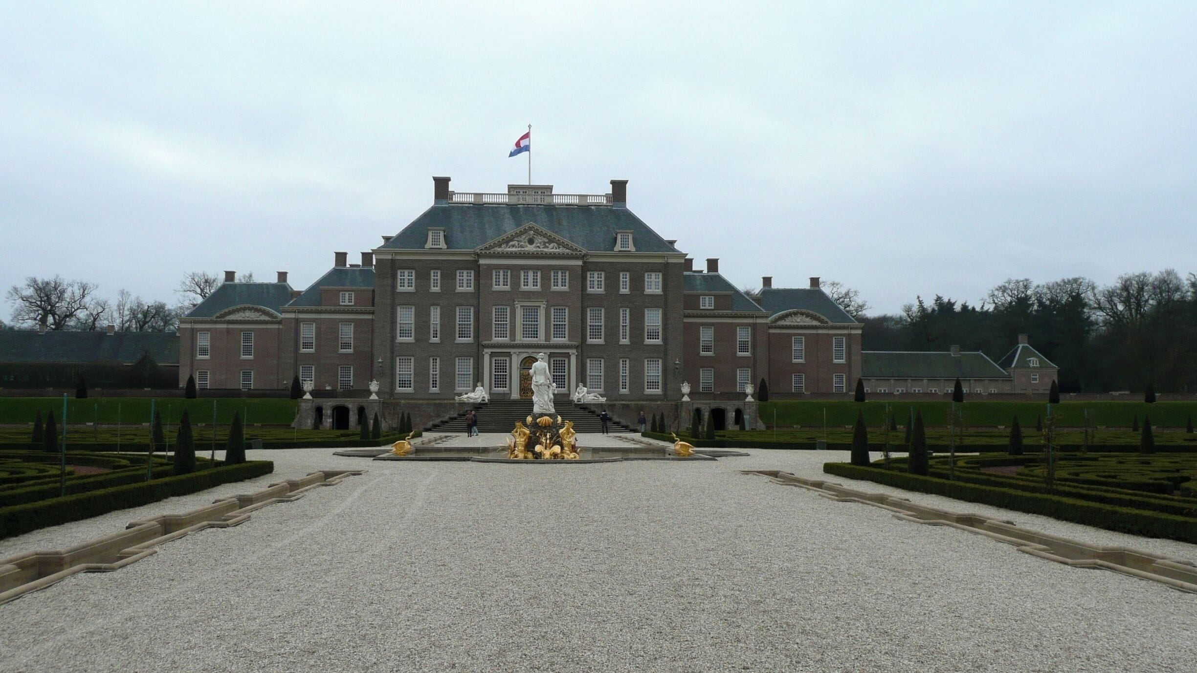 A large stately building with a symmetrical facade, a central statue, and a formal garden with geometric hedges.