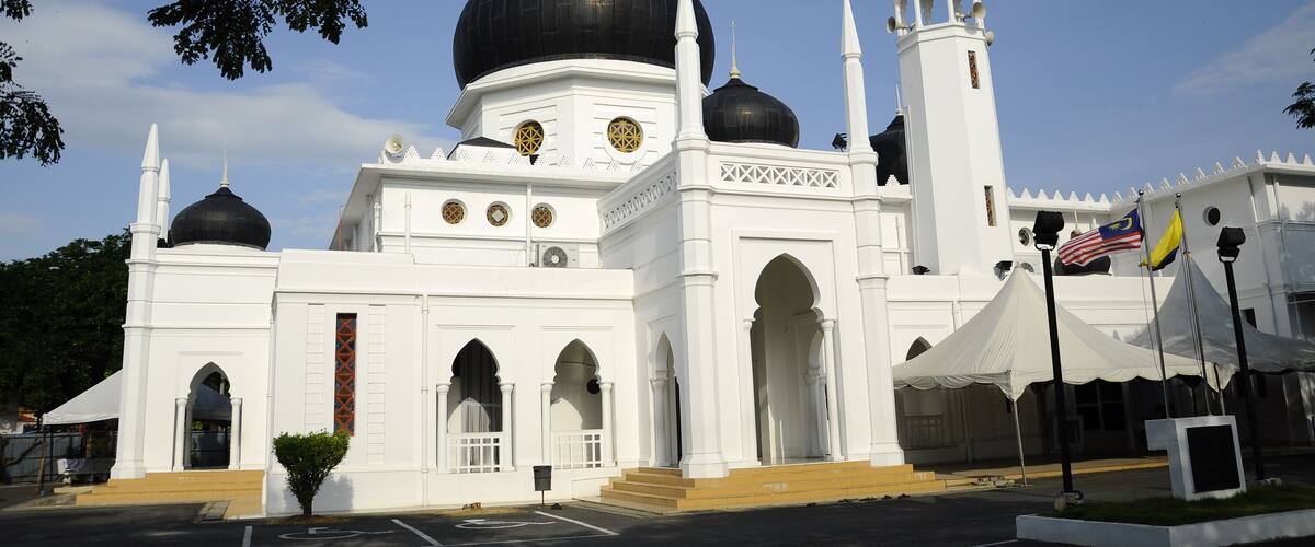A white mosque with multiple domes and minarets, a flagpole with flags, and a clear blue sky.