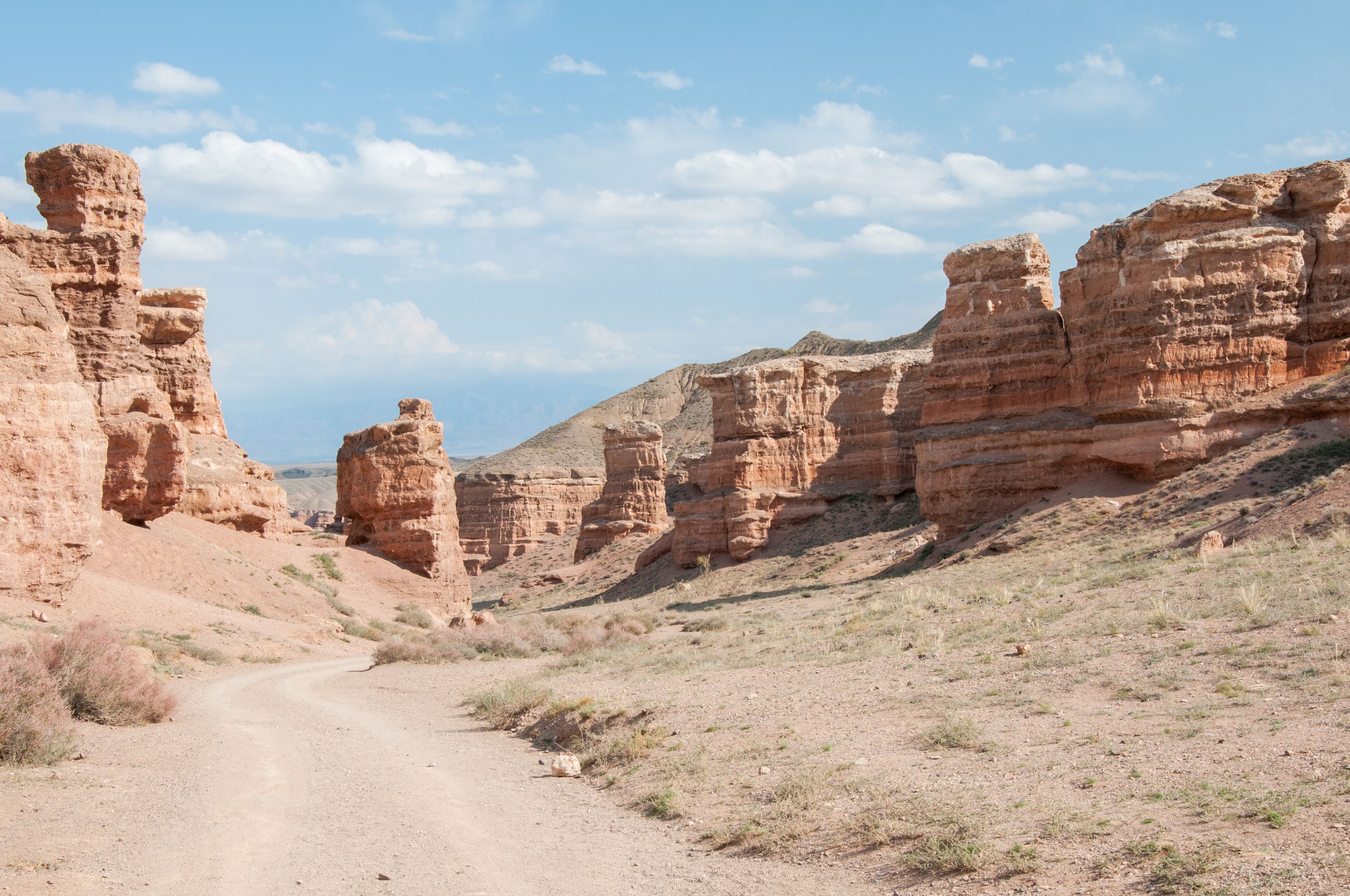 A desert landscape with a dirt road leading through a canyon with layered rock formations.