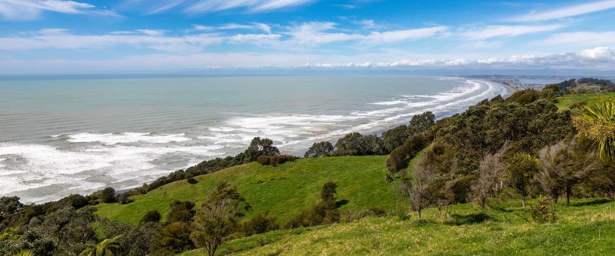 View of Ohope Beach with rolling green hills, crashing waves, and wispy clouds in the sky in Ohope, Bay of Plenty Region, New Zealand.