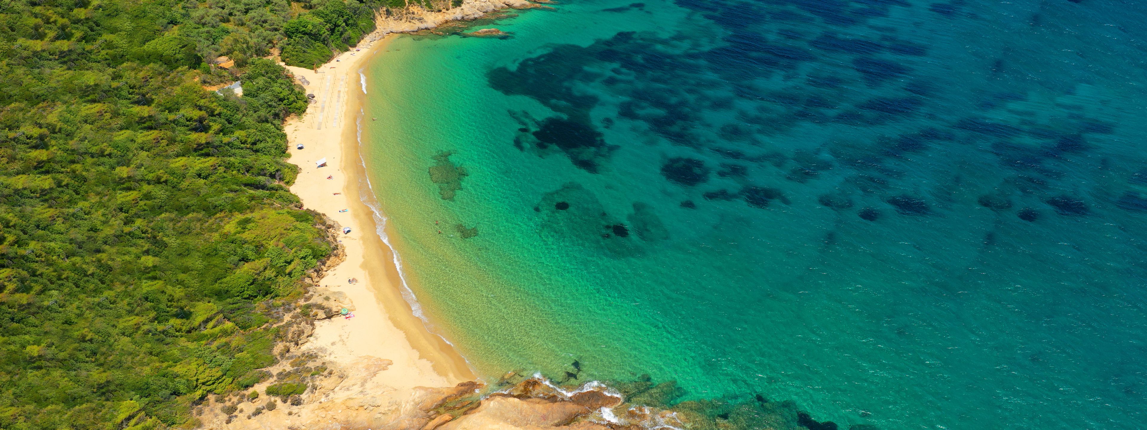 An aerial view of a beach with clear turquoise waters, sandy shore, and dense green vegetation.