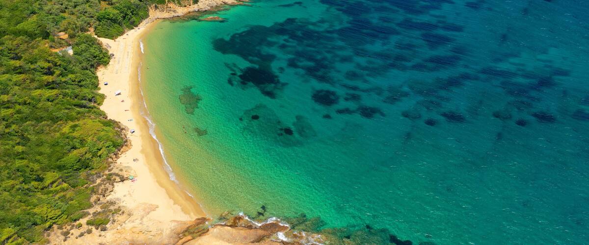 An aerial view of a beach with clear turquoise waters, sandy shore, and dense green vegetation.