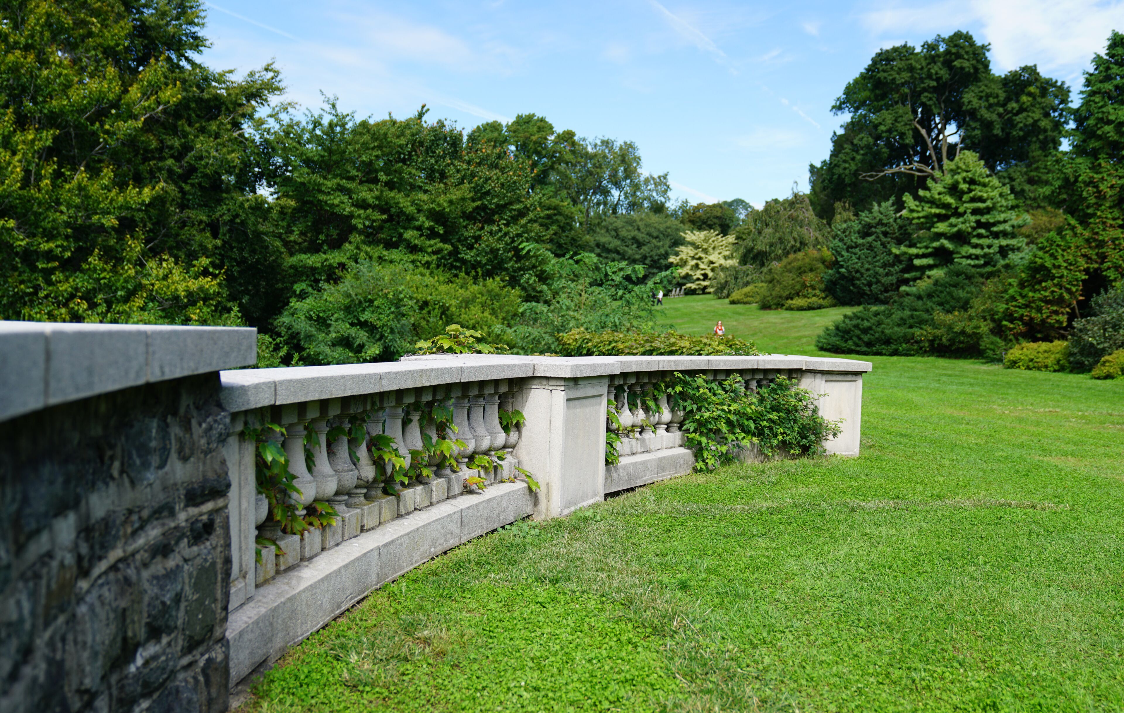 A stone balustrade with climbing plants, a well-maintained lawn, and dense trees in the background.