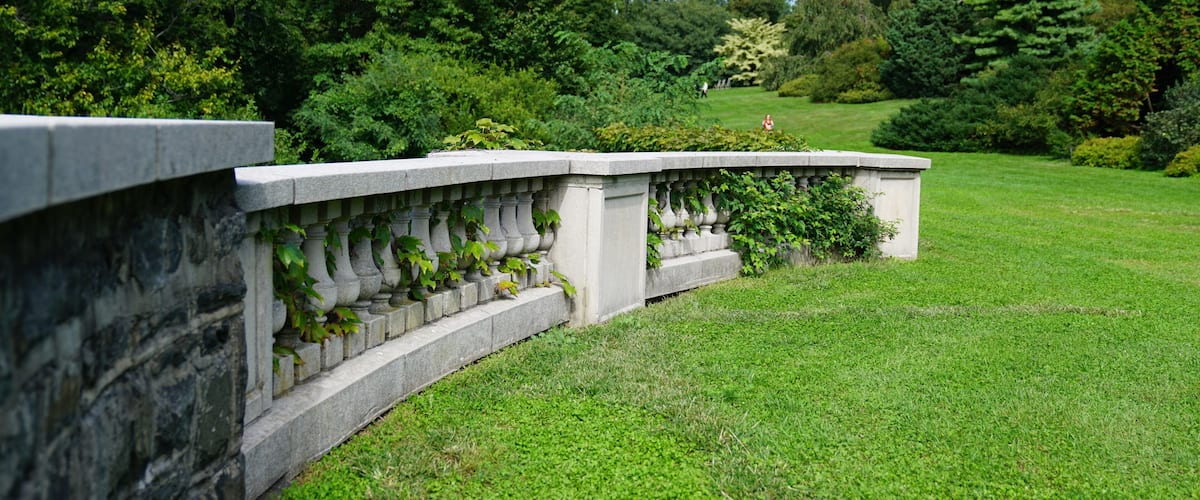 A stone balustrade with climbing plants, a well-maintained lawn, and dense trees in the background.