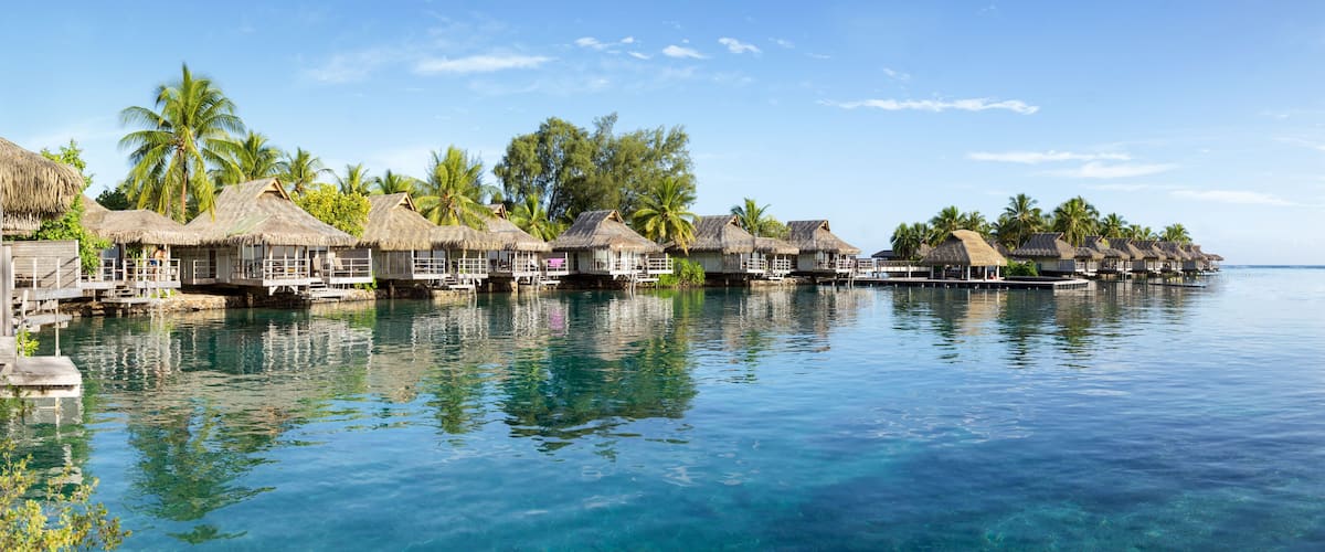 Overwater bungalows with thatched roofs, palm trees, and clear blue water.
