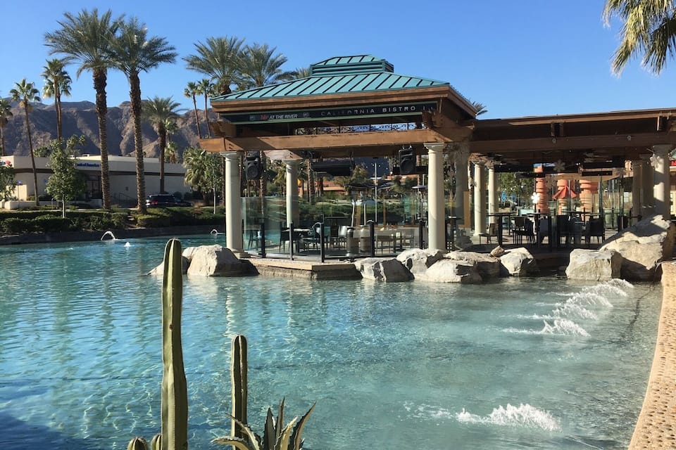 A pool with clear water, surrounded by palm trees and a cactus planter. In the background, there is a building with a green roof and columns.