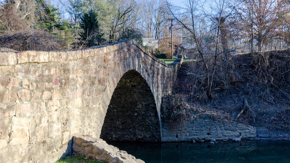 A stone bridge over a river with trees and a building in the background.
