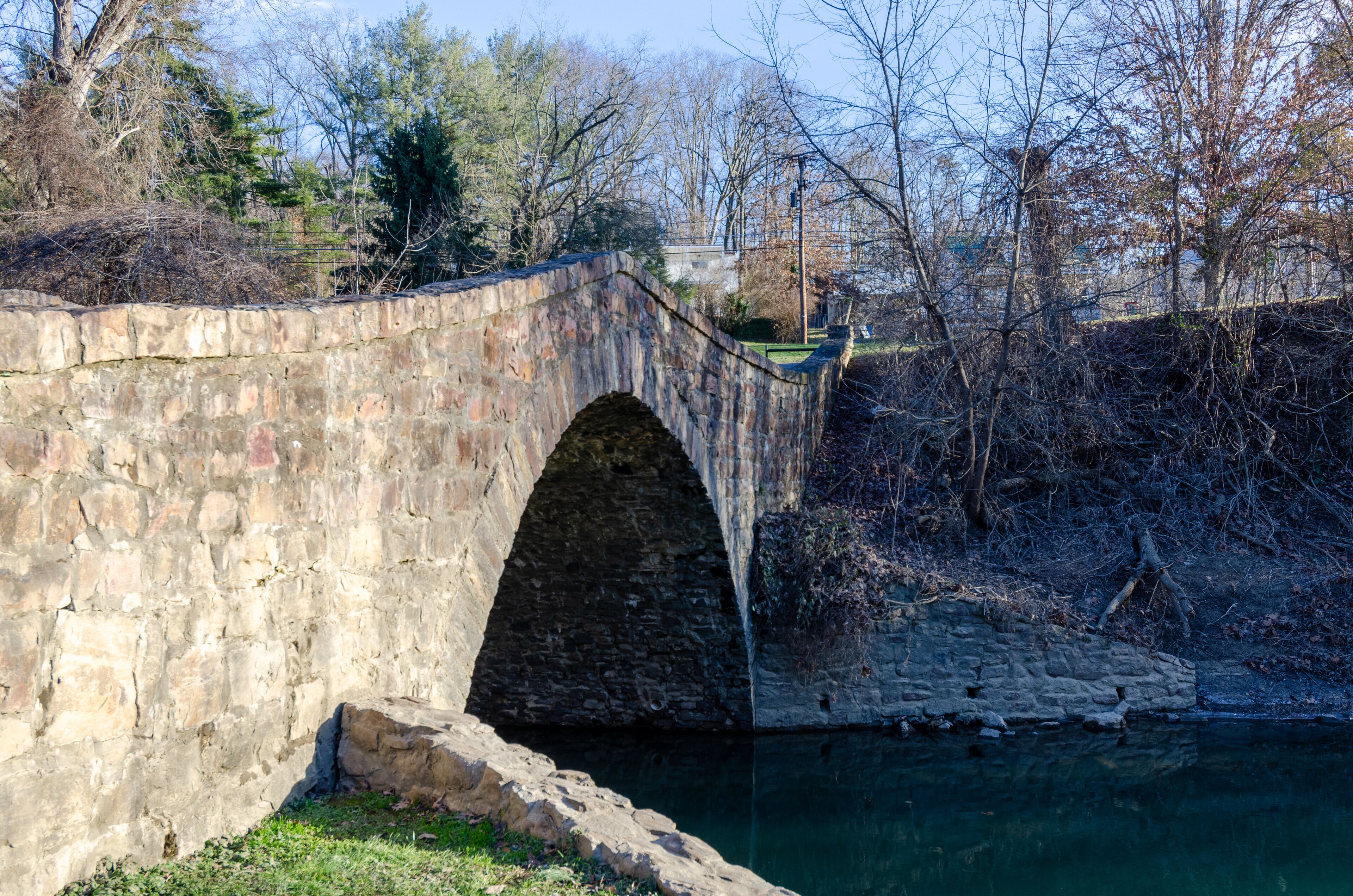 A stone bridge over a river with trees and a building in the background.