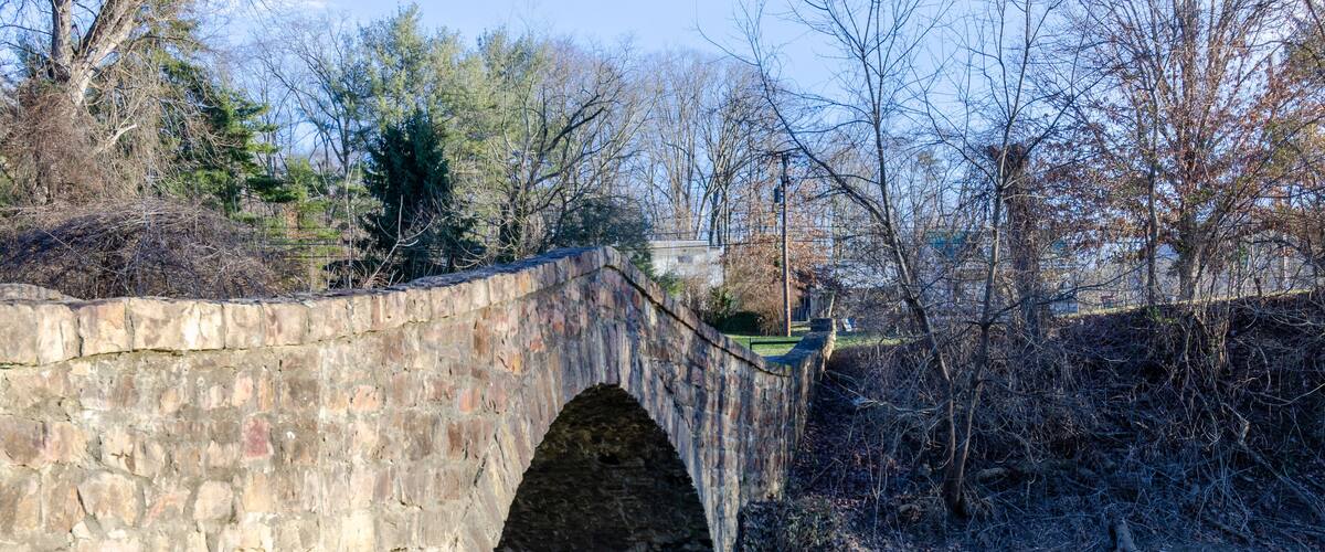 A stone bridge over a river with trees and a building in the background.