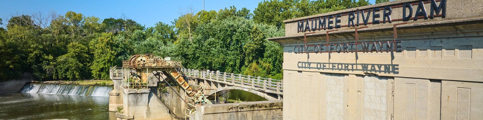 Sunny Maumee River Dam City of Fort Wayne sign on building with basin and waterfalls beneath green trees aerial