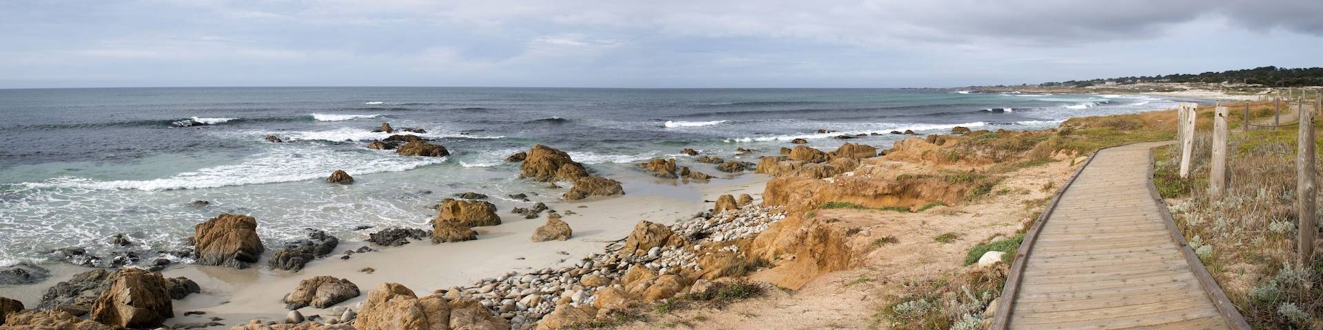 A coastal landscape with a wooden boardwalk, rocky shoreline, and the ocean.