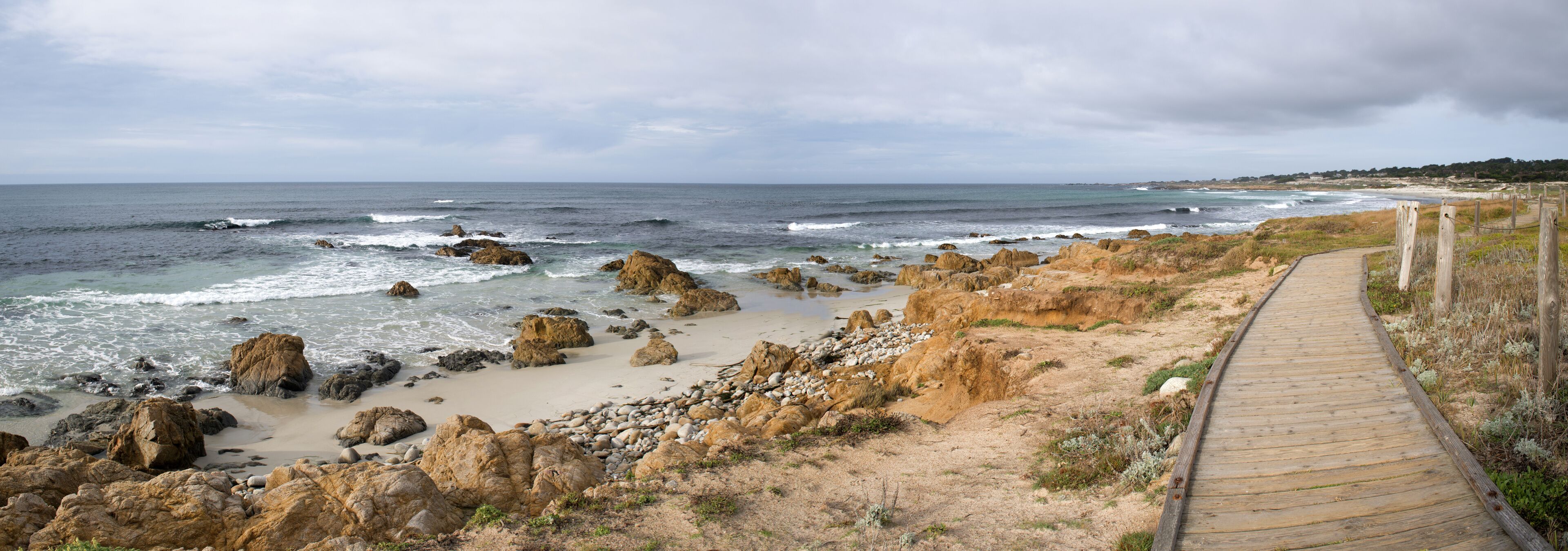 A coastal landscape with a wooden boardwalk, rocky shoreline, and the ocean.