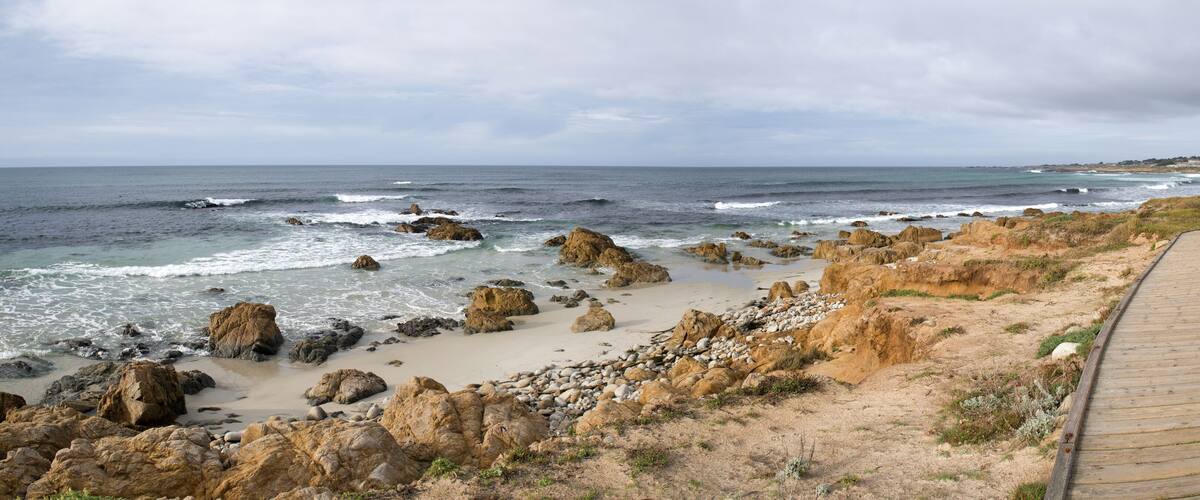 A coastal landscape with a wooden boardwalk, rocky shoreline, and the ocean.