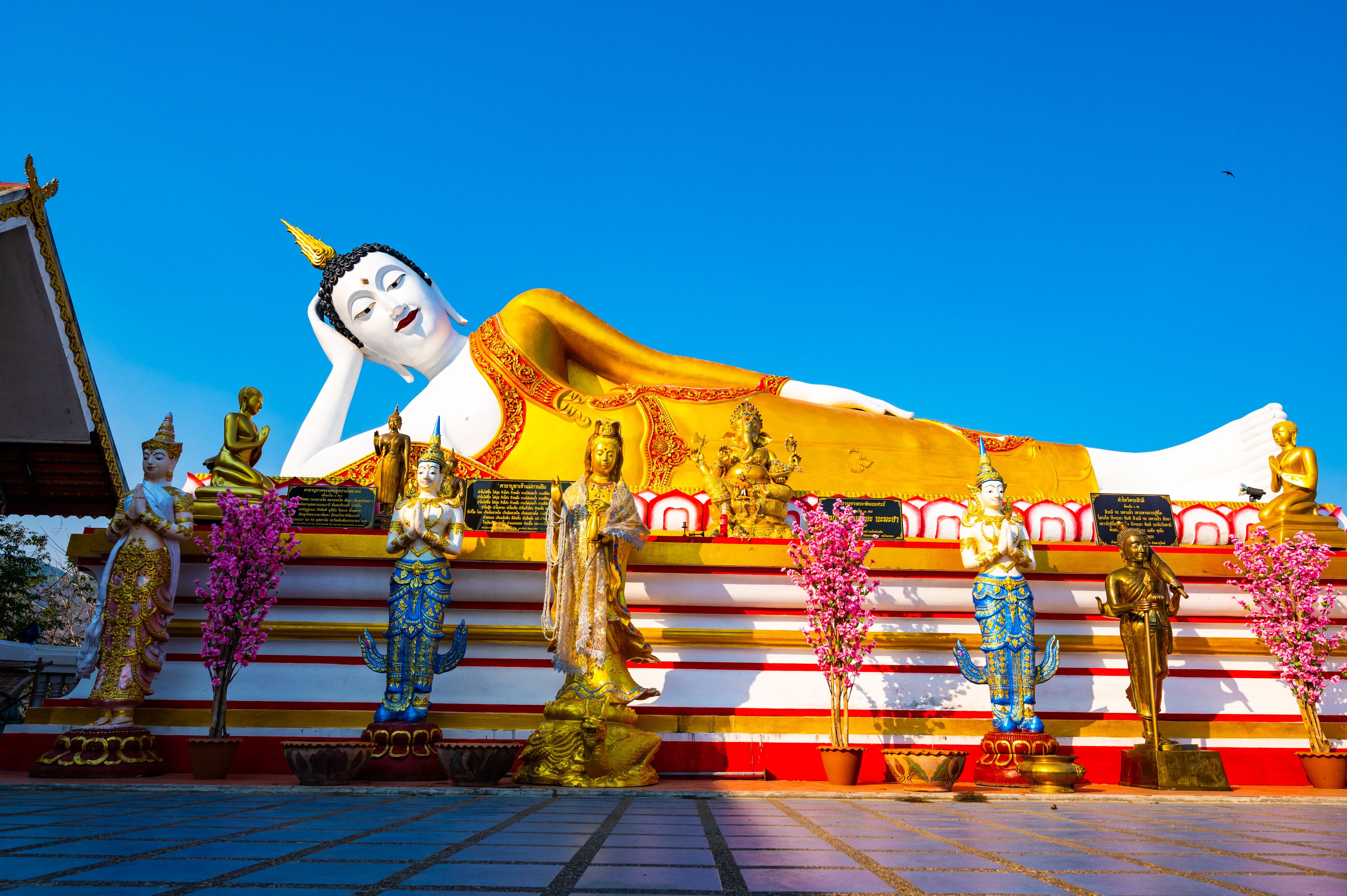 A large reclining Buddha statue with golden and white colors, surrounded by smaller statues and flowers.