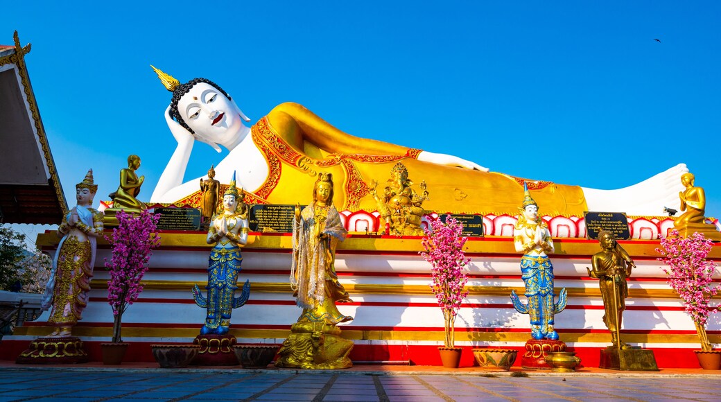 A large reclining Buddha statue with golden and white colors, surrounded by smaller statues and flowers.
