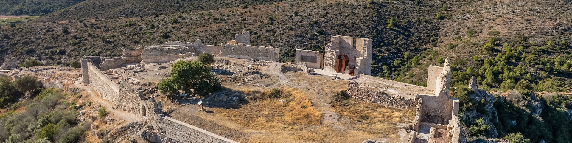 Une ancienne forteresse de pierre, entourée de prairies arides et de quelques arbres clairsemés, avec des montagnes en arrière-plan.
