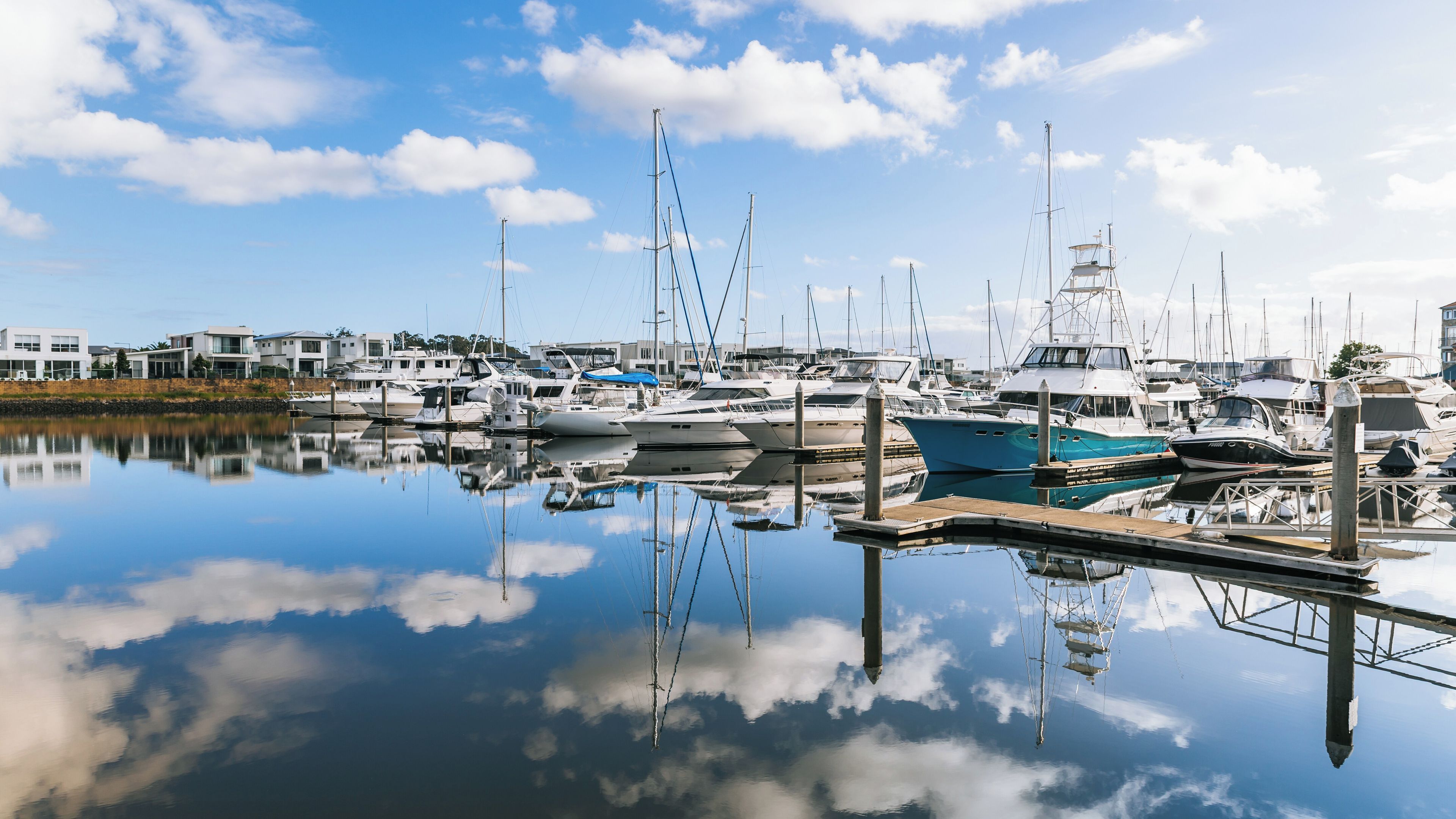 A marina with various boats docked, a calm water surface reflecting the sky and boats, and modern buildings in the background.