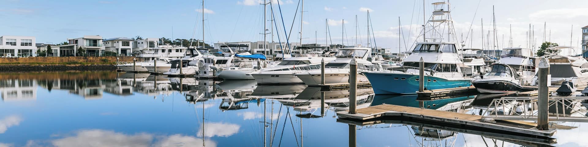 A marina with various boats docked, a calm water surface reflecting the sky and boats, and modern buildings in the background.