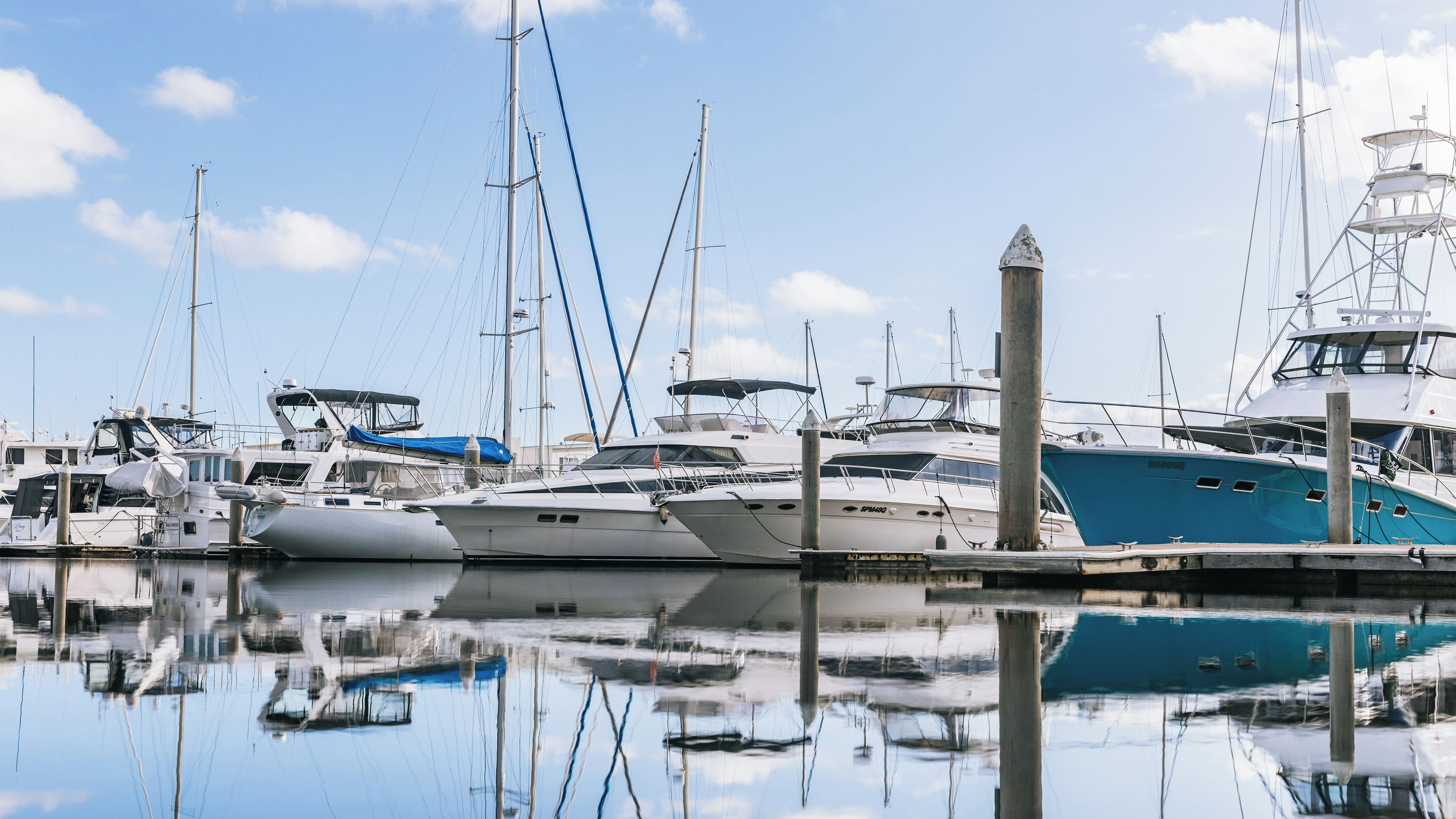 A marina with multiple yachts docked, calm water reflecting the boats, and a clear sky.