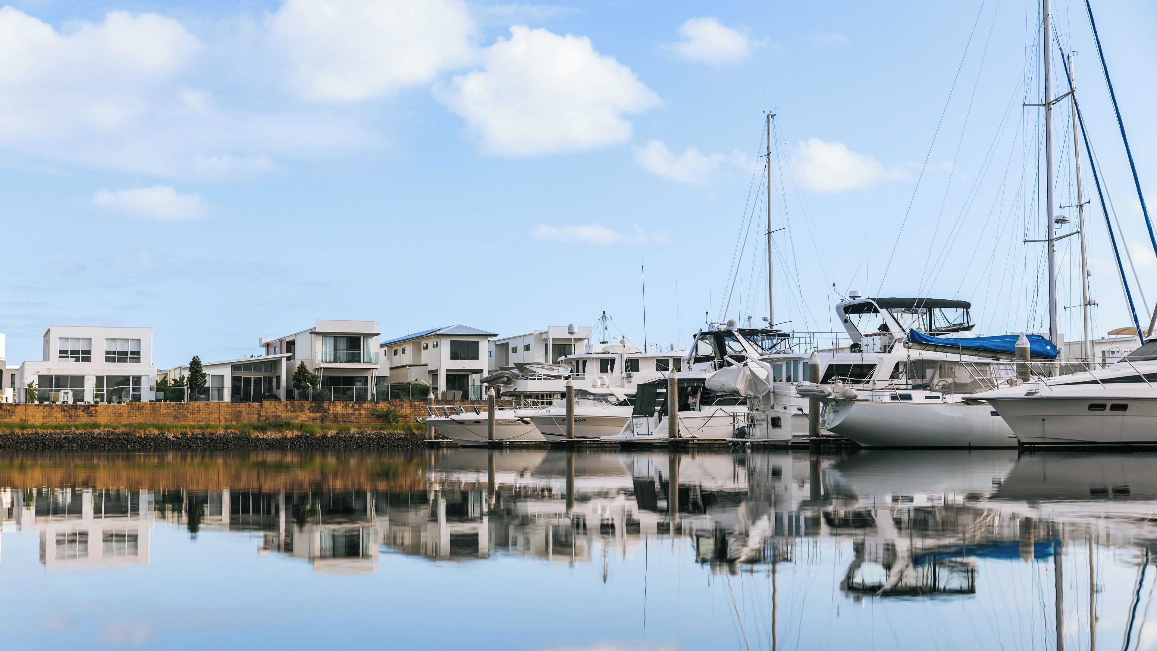 A marina with yachts docked, a calm body of water, and modern buildings in the background.