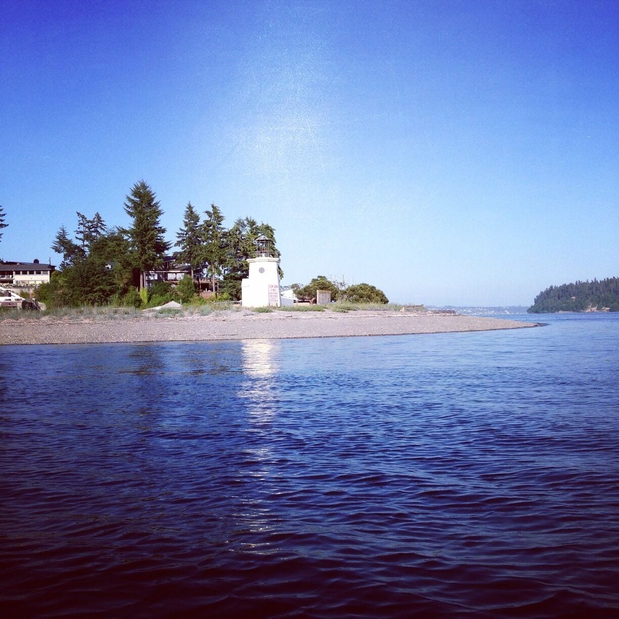 A calm lake with a small white lighthouse on a sandy shore, surrounded by trees and buildings.