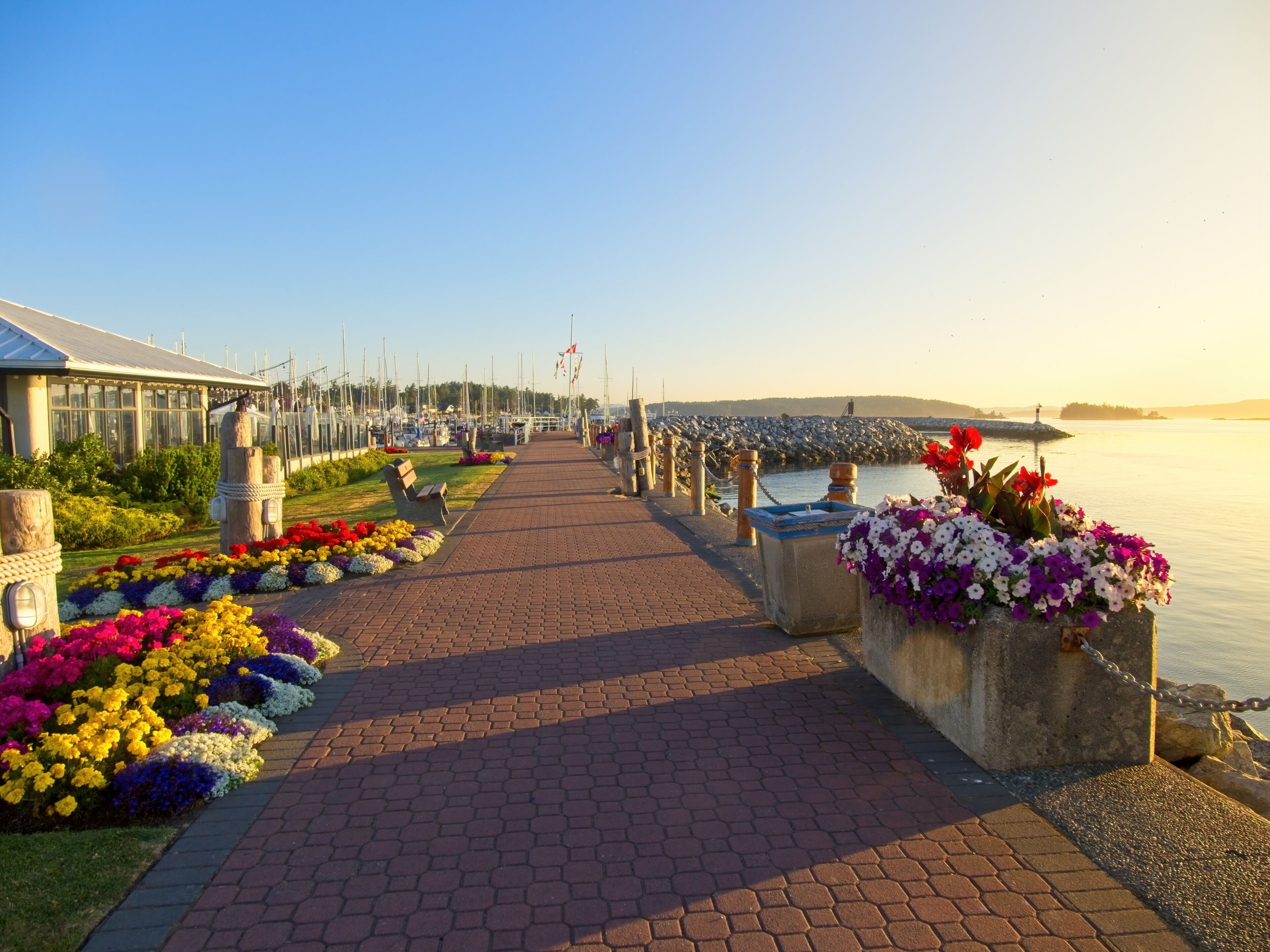 A waterfront promenade with a brick pathway, flower beds, and a marina in the background.