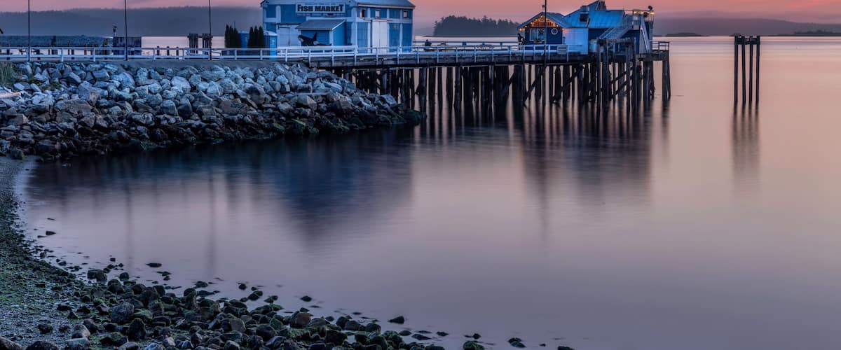 Une jetée s’avance dans des eaux calmes, avec un bâtiment bleu portant l’inscription « From Market » et une construction plus petite surmontée d’un toit jaune.