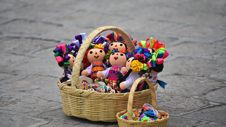 A wicker basket filled with colorful dolls and a smaller basket with more dolls on a paved surface.