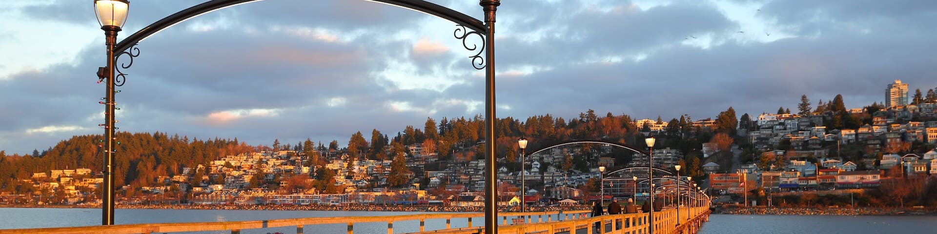 A wooden pier with street lamps extending over a body of water, leading to a cityscape with buildings and trees in the background.